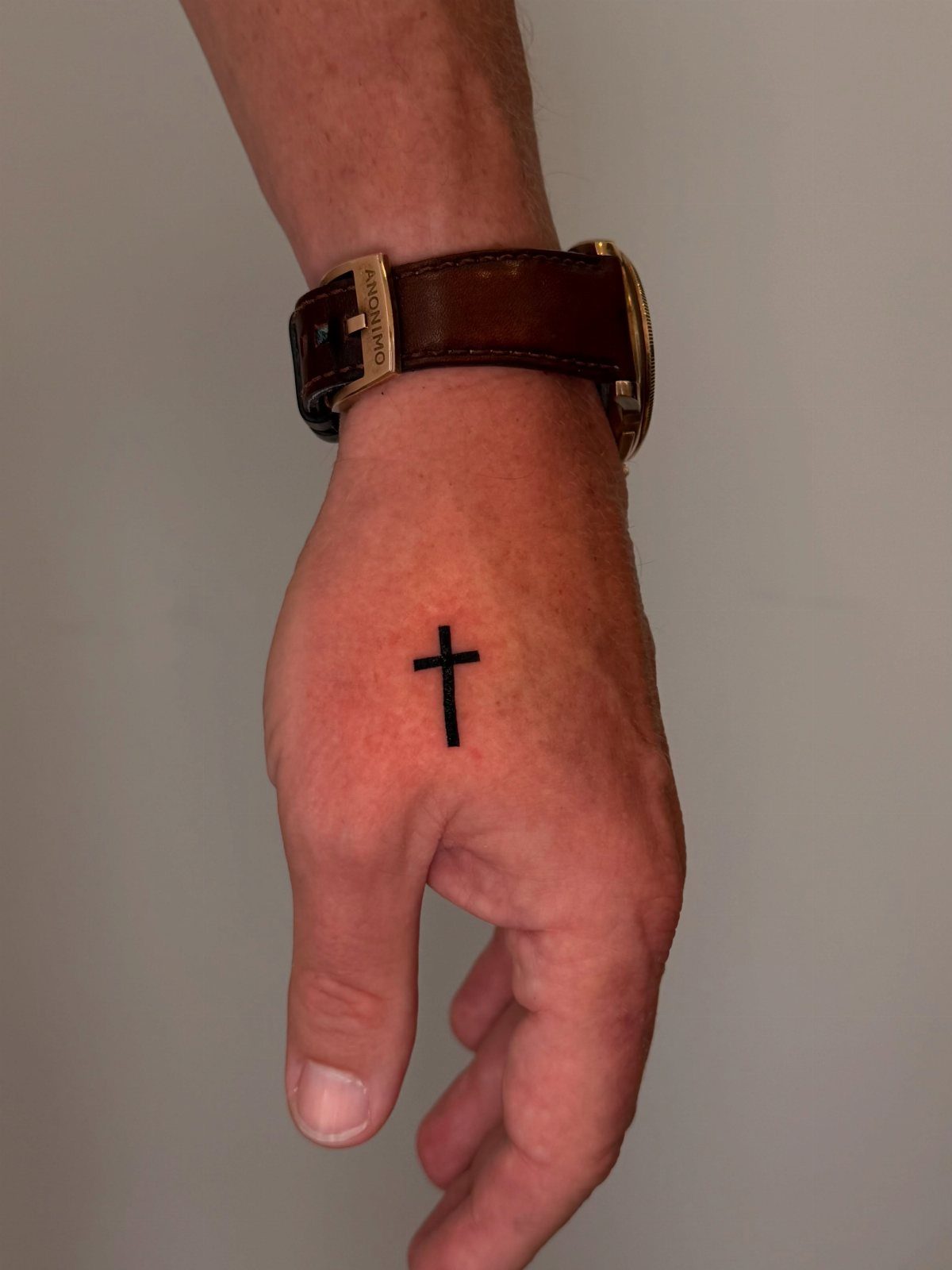 Small black cross tattoo on the back of a man's hand wearing a watch