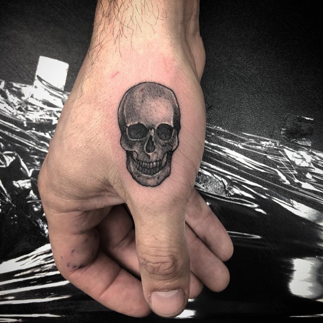 Small skull tattoo on the back of a man's hand in black and grey ink.
