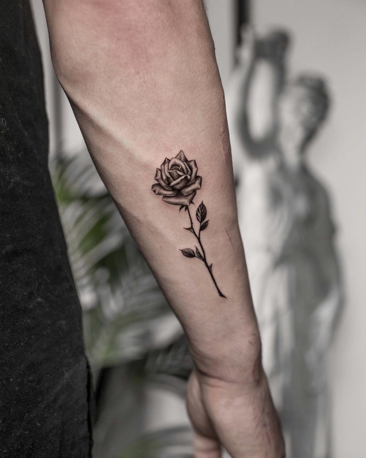 Small black ink rose tattoo on a person's forearm with a detailed stem.