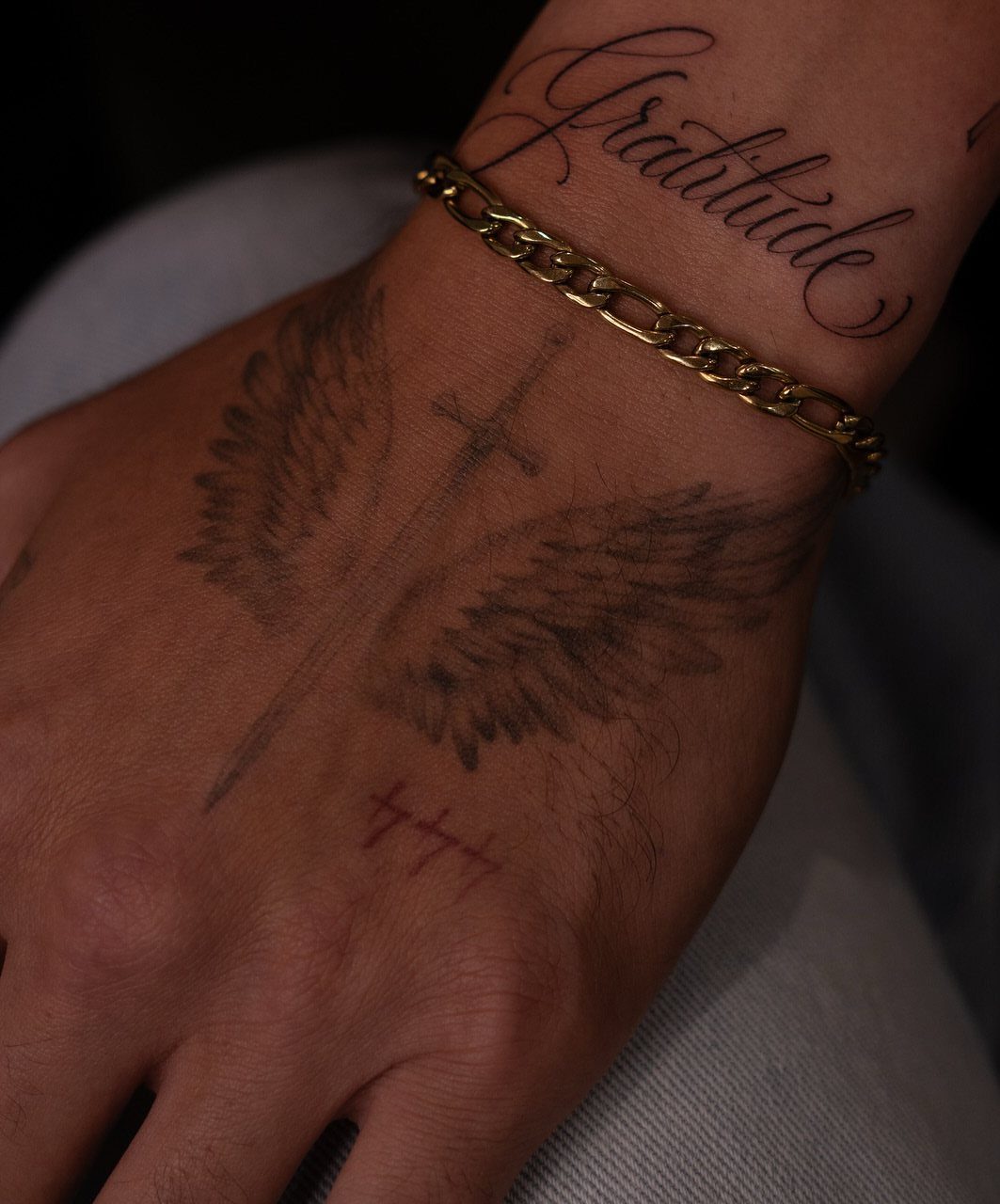 Cursive Gratitude tattoo on a wrist next to a gold chain bracelet.