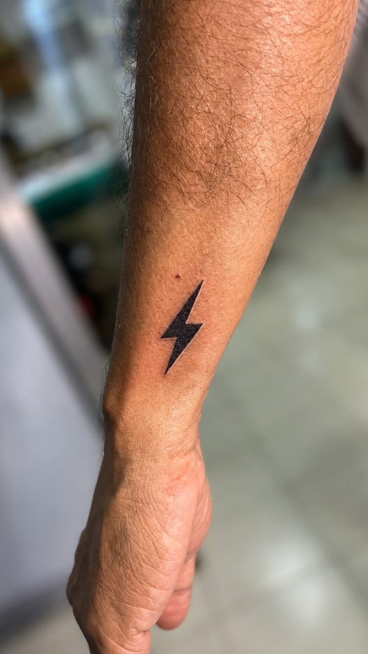 Small black lightning bolt tattoo on the inner wrist of a hairy arm.