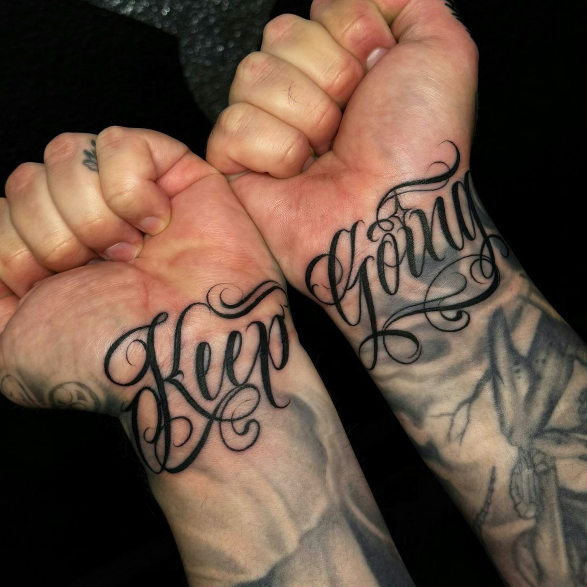 Cursive black ink tattoos on inner wrists reading Keep Going on clenched fists.