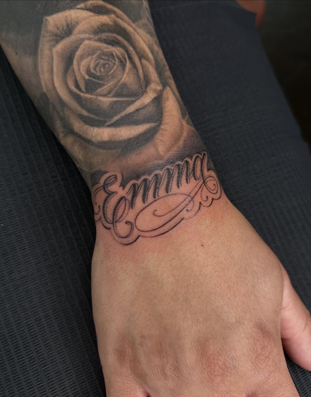 Cursive name tattoo 'Emma' on a wrist below a large grey-scale rose tattoo.