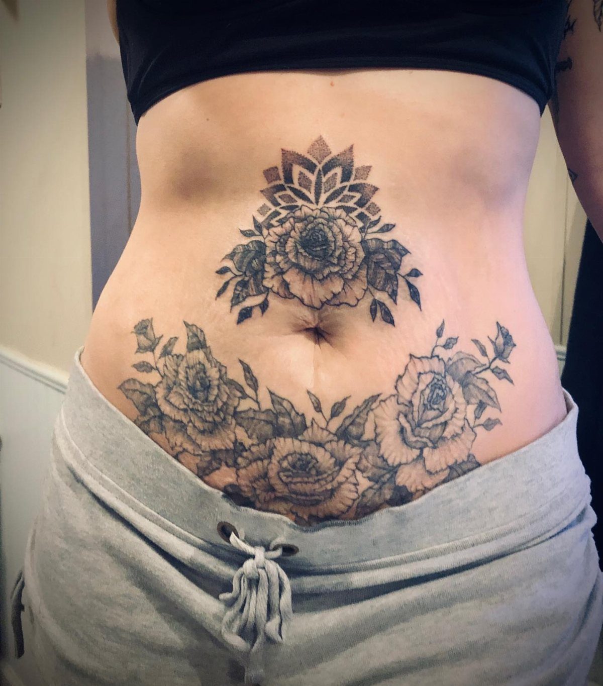 Woman's stomach with black ink rose and mandala tattoos around the belly button.