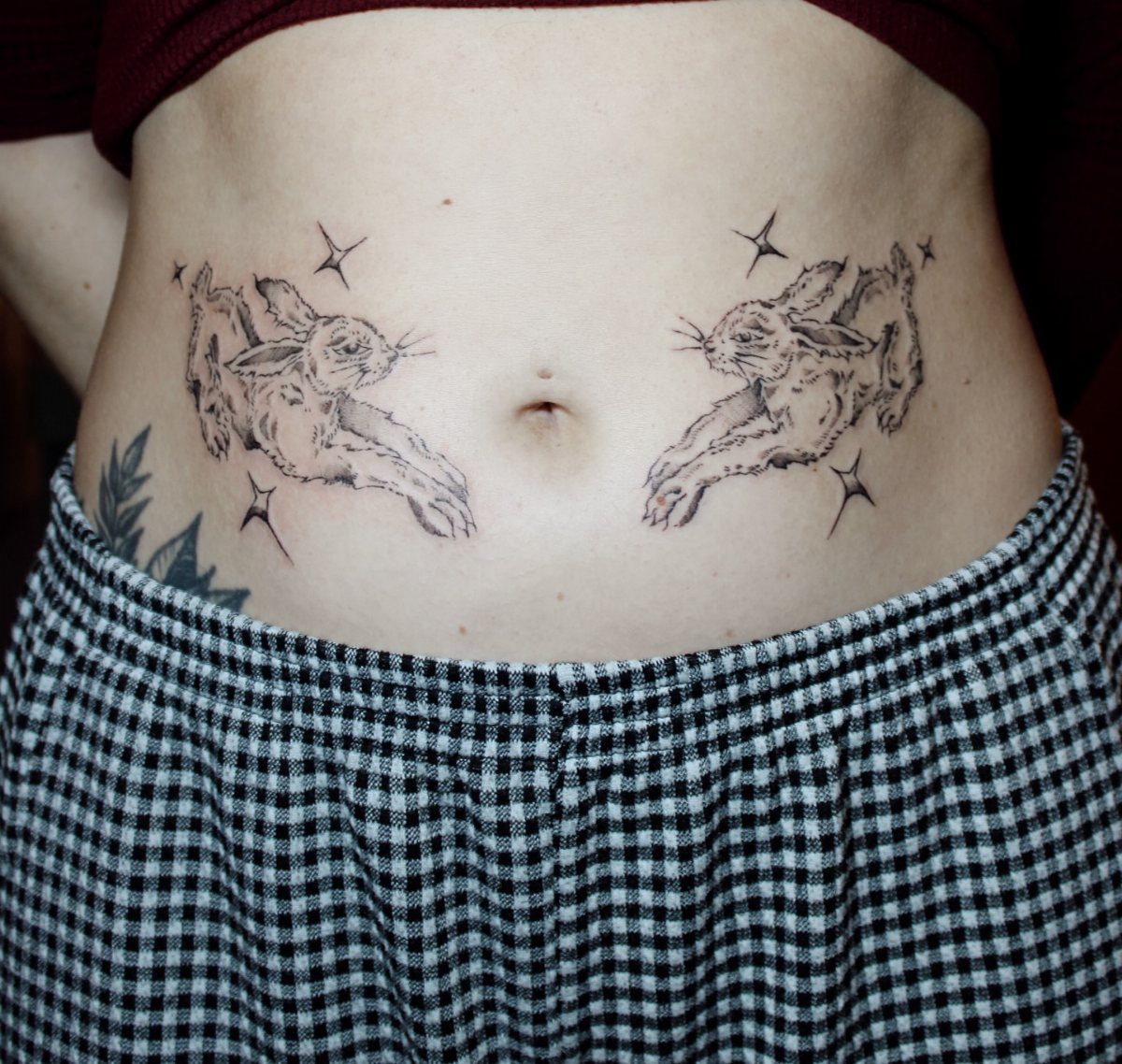 Two black and gray rabbit tattoos on a woman's stomach with small stars.