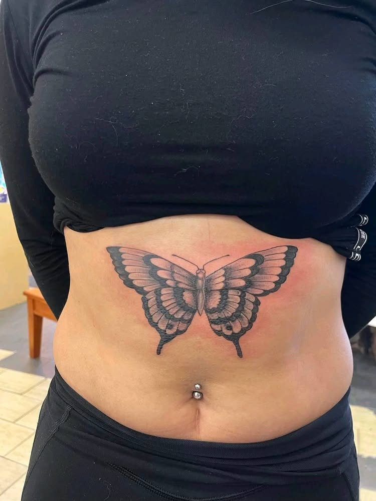 Butterfly tattoo on a woman's stomach with a belly button piercing and black clothing