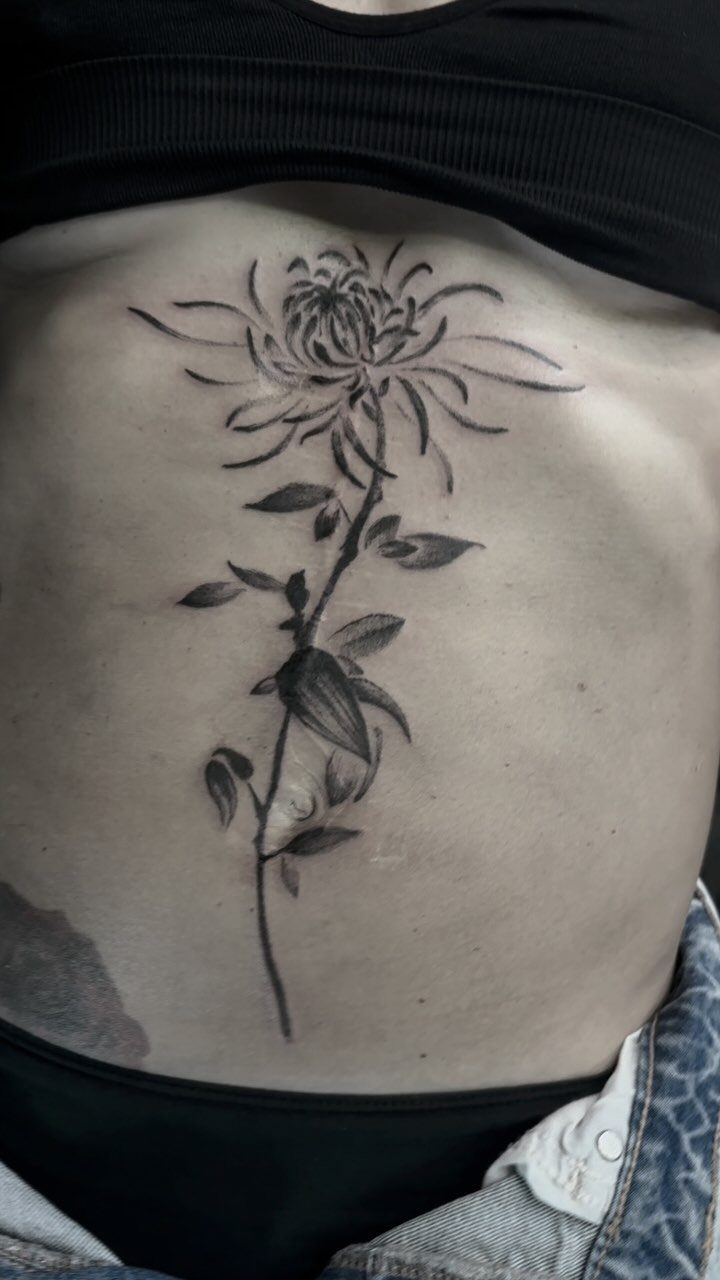 Black ink flower tattoo with a long stem and leaves on a stomach.
