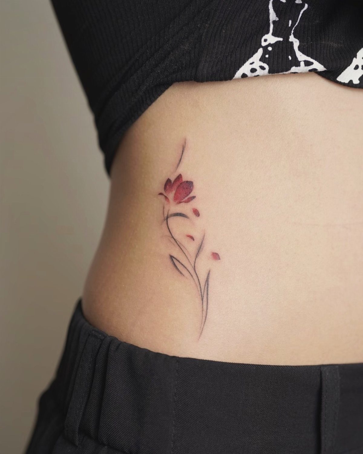 Small red flower tattoo with a thin stem on a woman's stomach.