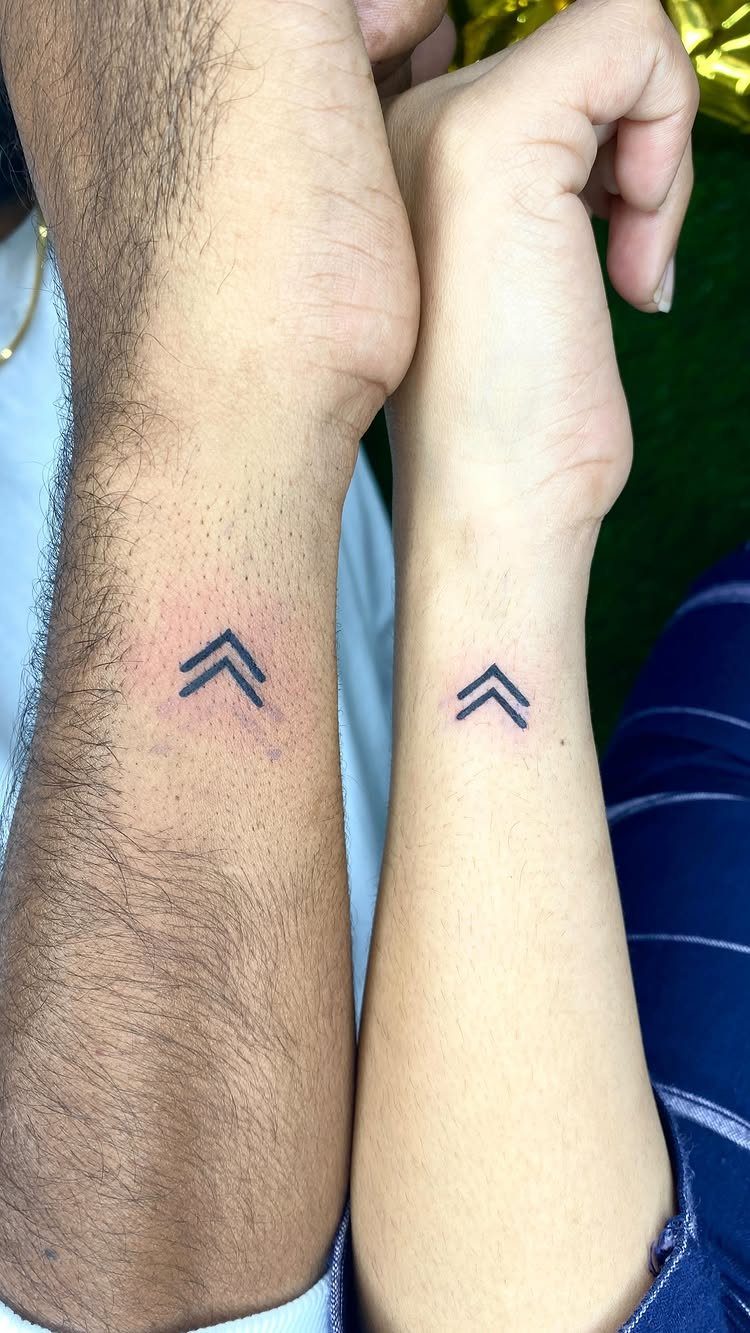 Two wrists with matching small black arrow tattoos pointing upwards.