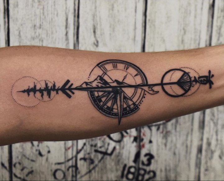 Forearm tattoo featuring a compass, clock, mountains, and an arrow in black ink.