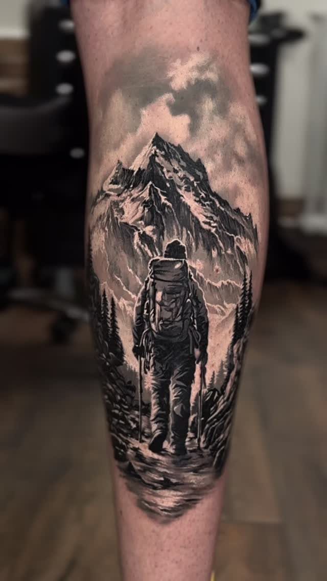 Hiker with backpack standing before a large snow-capped mountain peak tattoo on leg.