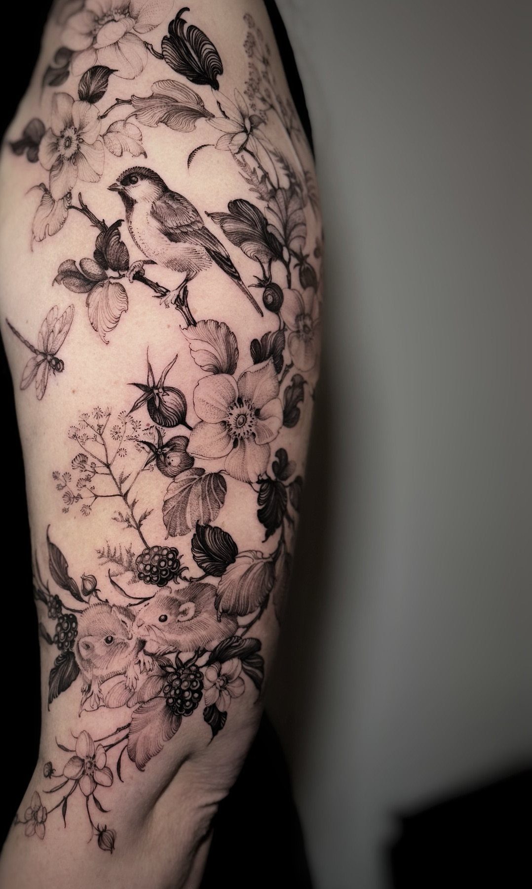 Detailed black ink nature tattoo on an arm featuring birds, flowers, and berries.