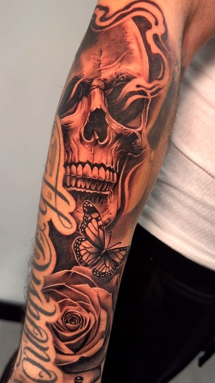 Detailed black and grey tattoo of a skull, butterfly, and rose on a forearm.