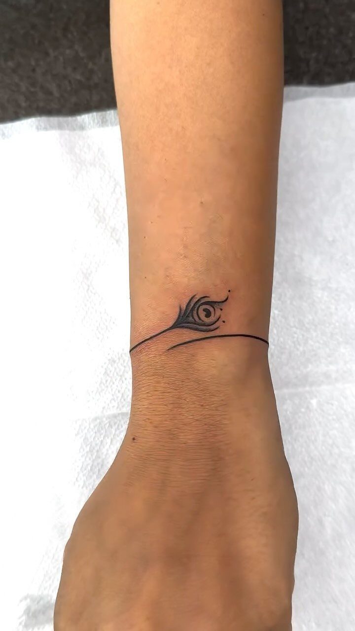 Small black feather tattoo wrapping around a person's ankle on a white background.