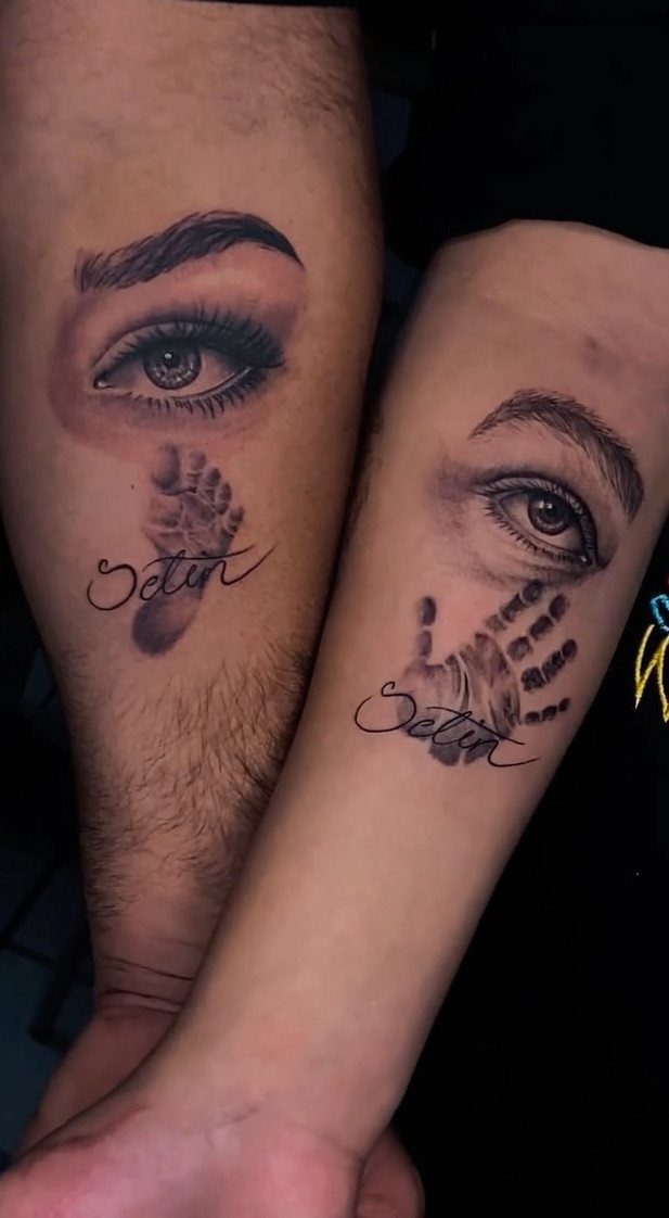 Tattoos of realistic eyes with a child's handprint and footprint on two arms.