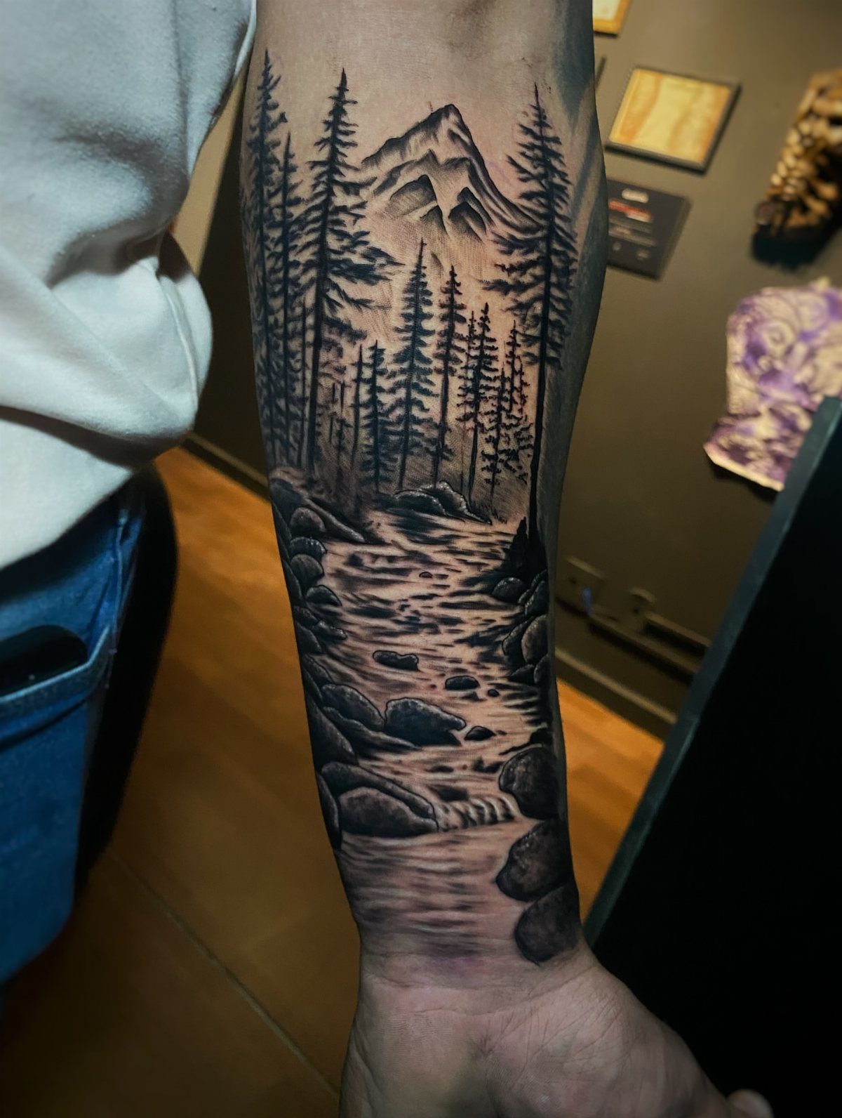 Mountain landscape tattoo on a man's forearm with pine trees and a river.