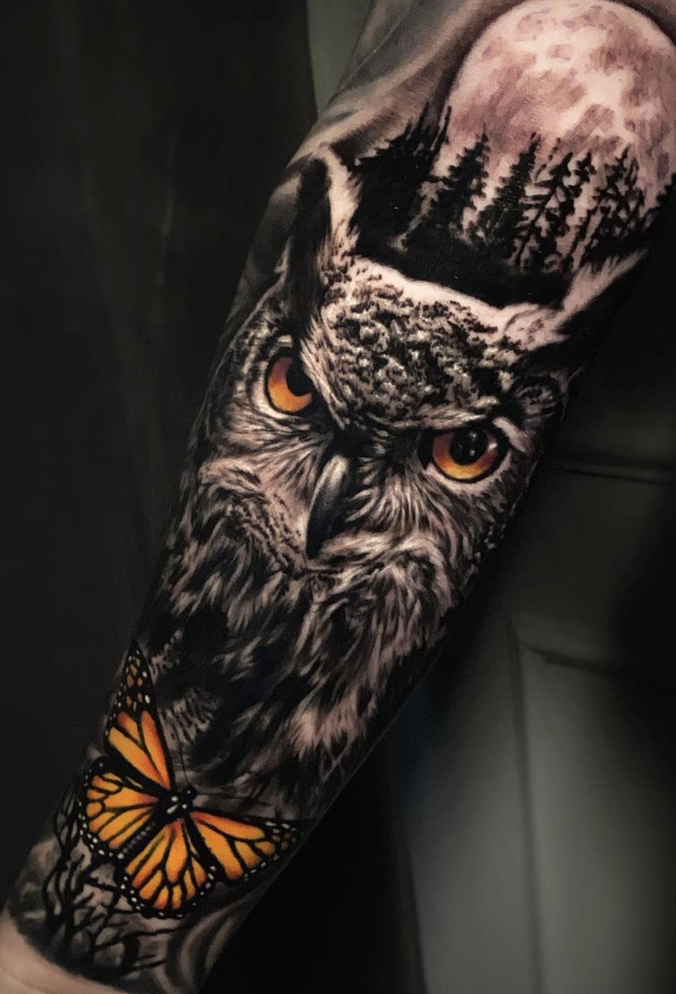 Detailed owl tattoo with orange eyes, a moon, and a butterfly on an arm.