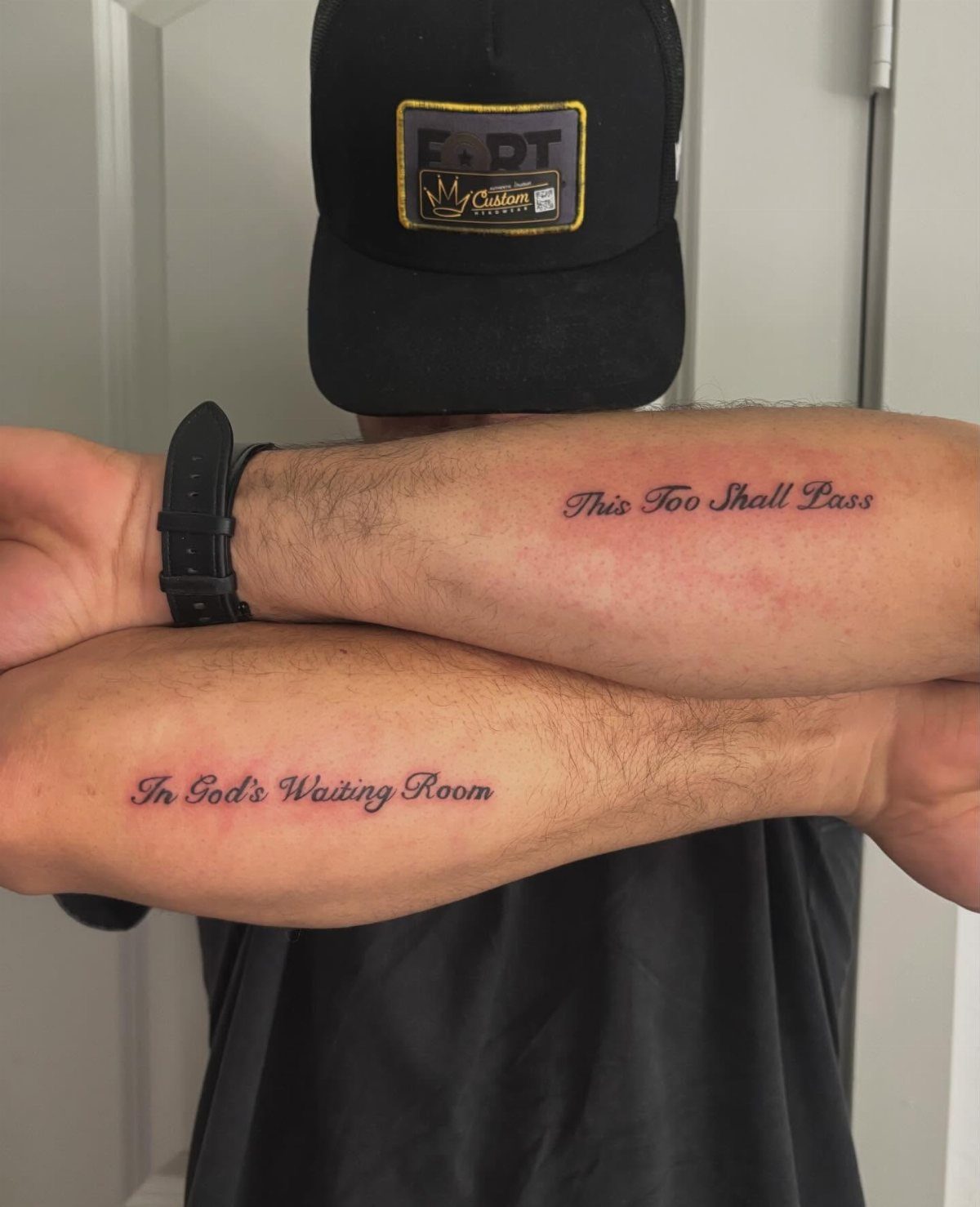 Crossed forearms with script tattoos reading This Too Shall Pass and In God's Waiting Room