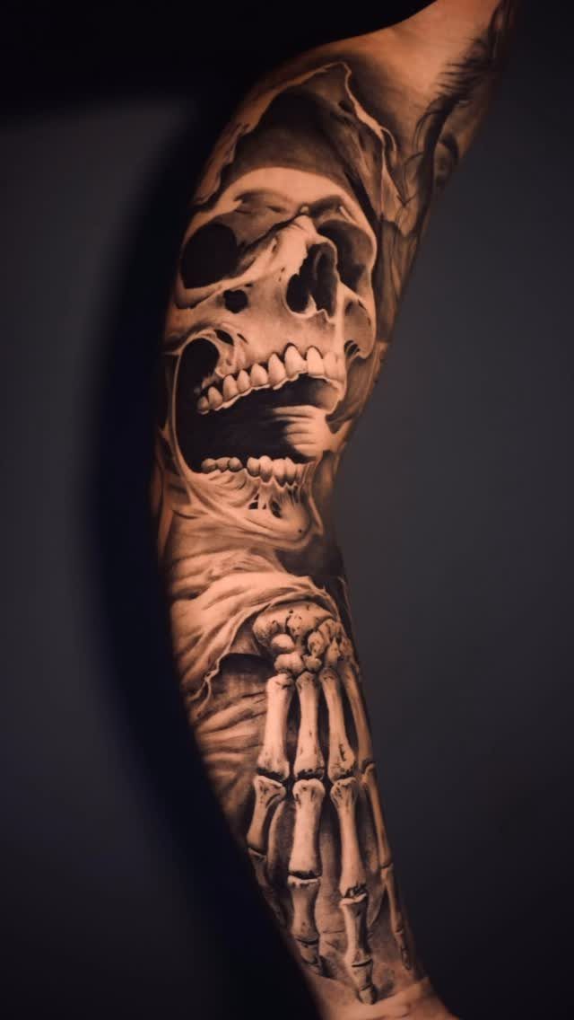 Screaming skull and skeletal hand tattoo in black and grey ink on an arm