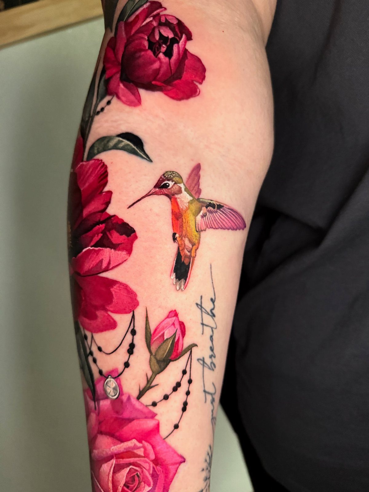 Hummingbird tattoo with red peonies and black beads on a person's arm