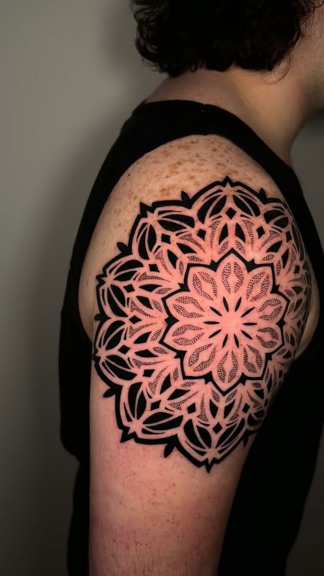 Symmetrical black ink mandala tattoo on a person's shoulder with intricate geometric patterns.