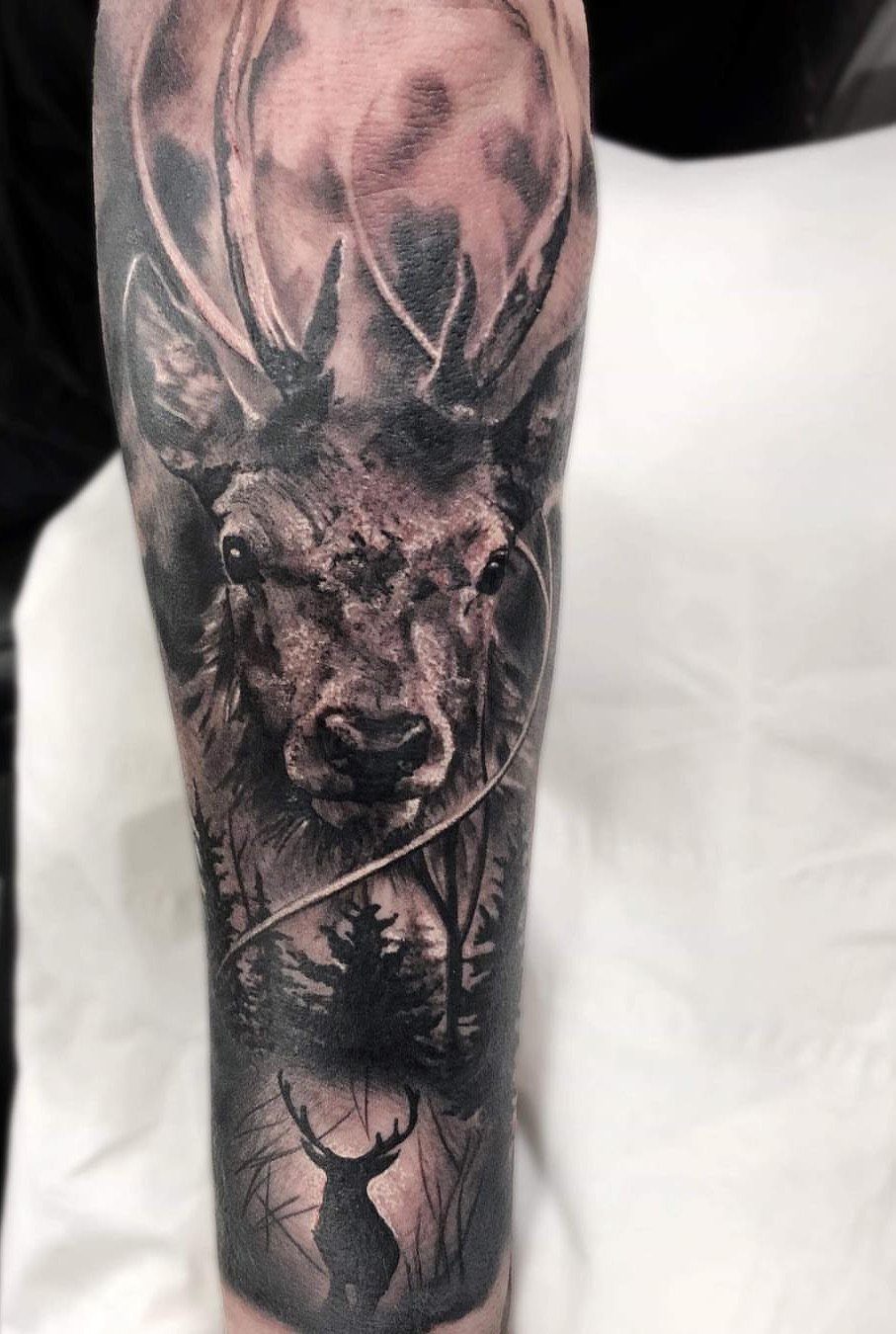 Realistic stag head tattoo with forest scenery in black and gray ink on arm