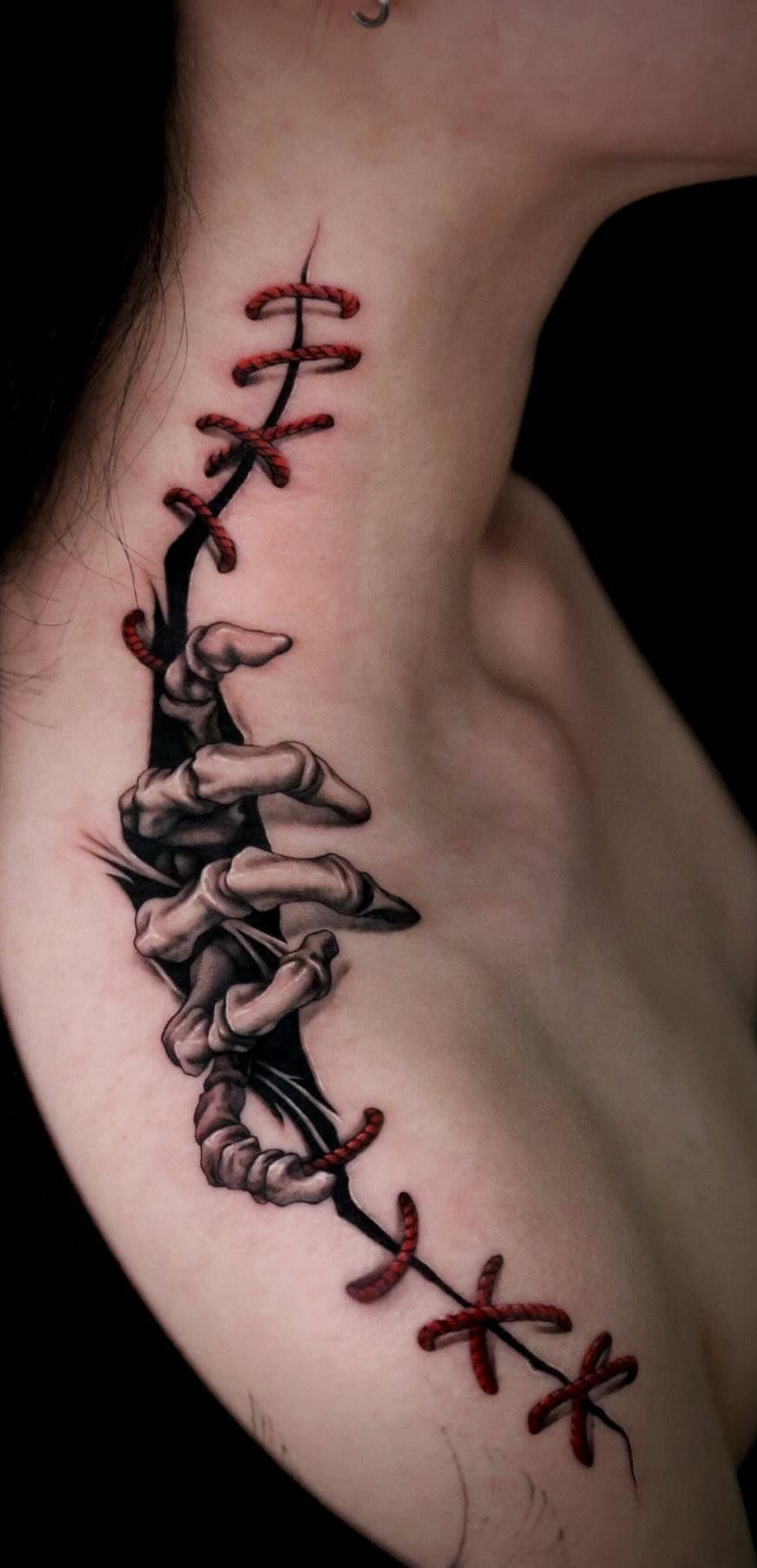 Skeletal hand tattoo emerging from skin with red stitching on a shoulder.