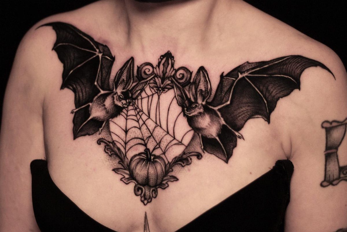Detailed black ink chest tattoo featuring two bats, a spiderweb, and a pumpkin.