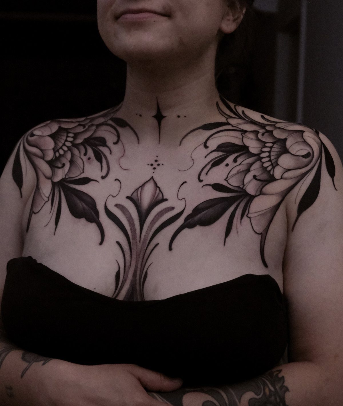 Symmetrical black and grey ornamental floral tattoo across a person's chest and collarbones