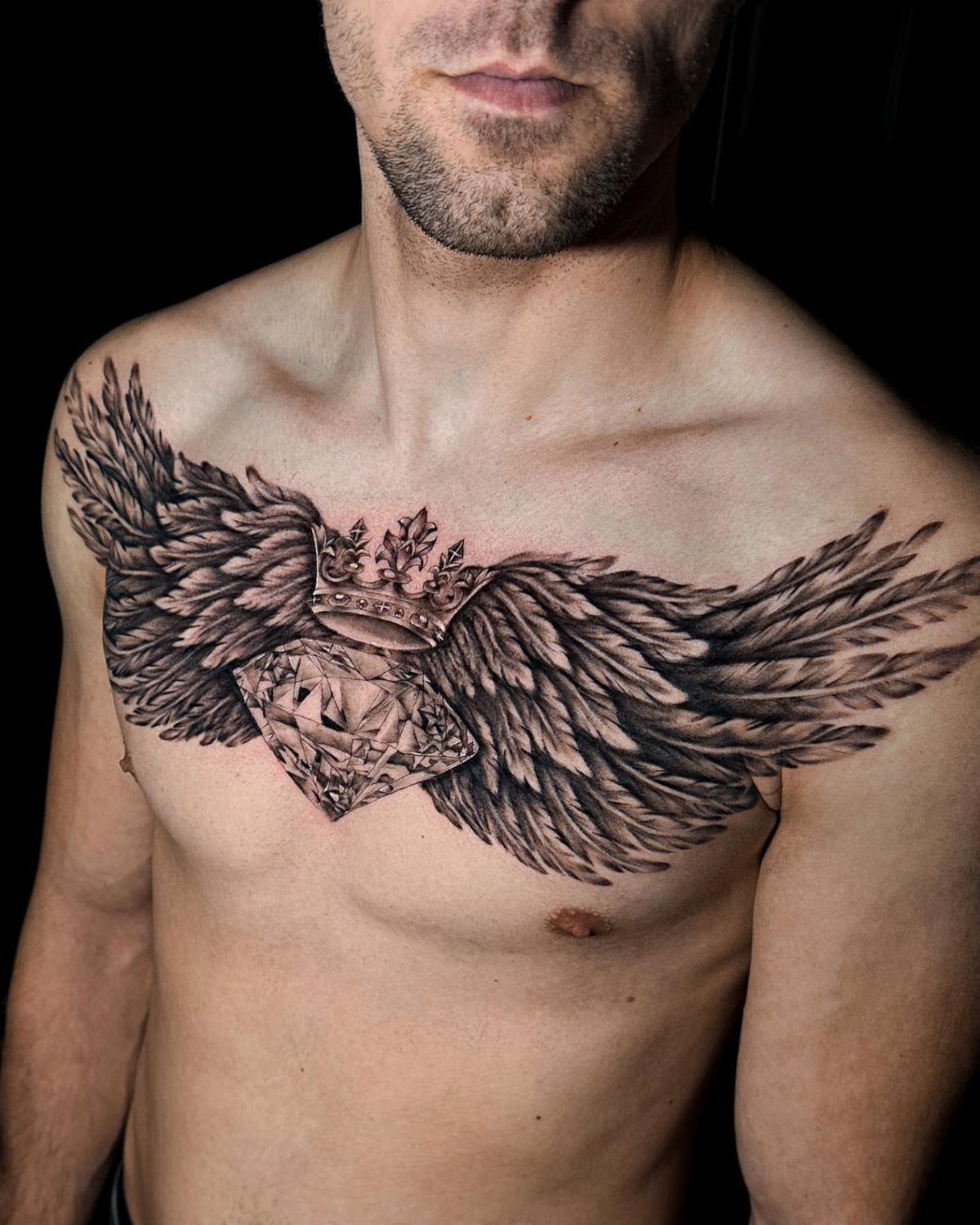 Detailed black and grey chest tattoo of a crowned diamond with large outstretched wings.