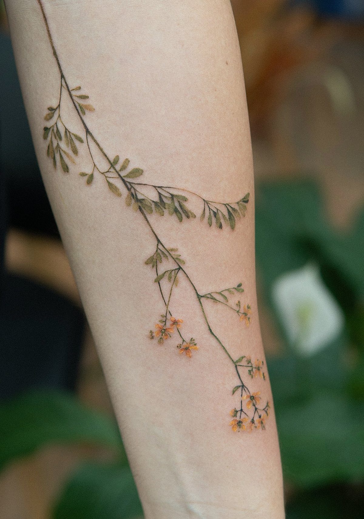 Botanical sprig tattoo with green leaves and orange flowers on a person's forearm.