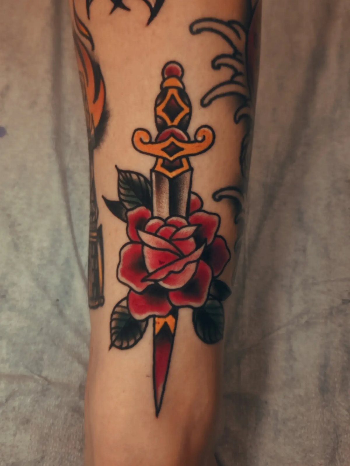 Traditional tattoo of a red rose and gold dagger on a person's arm.