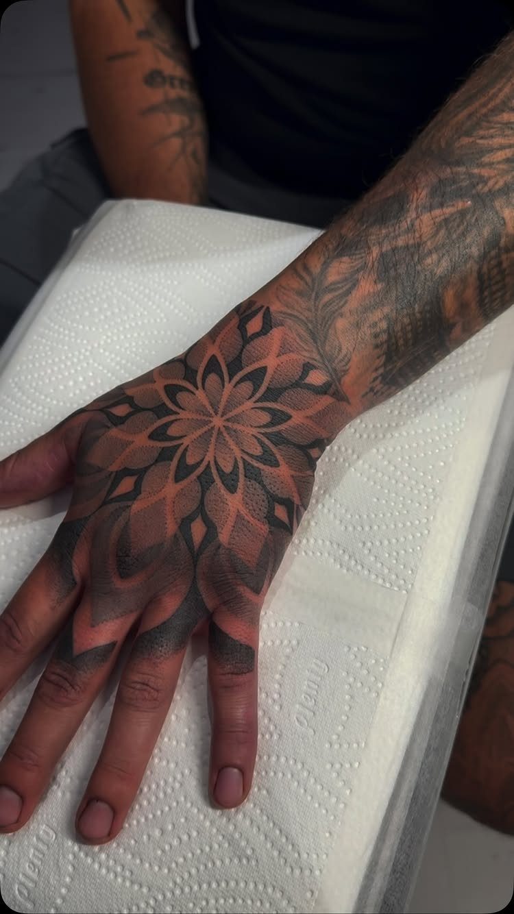 Man's hand with a detailed black and grey mandala tattoo on the back.