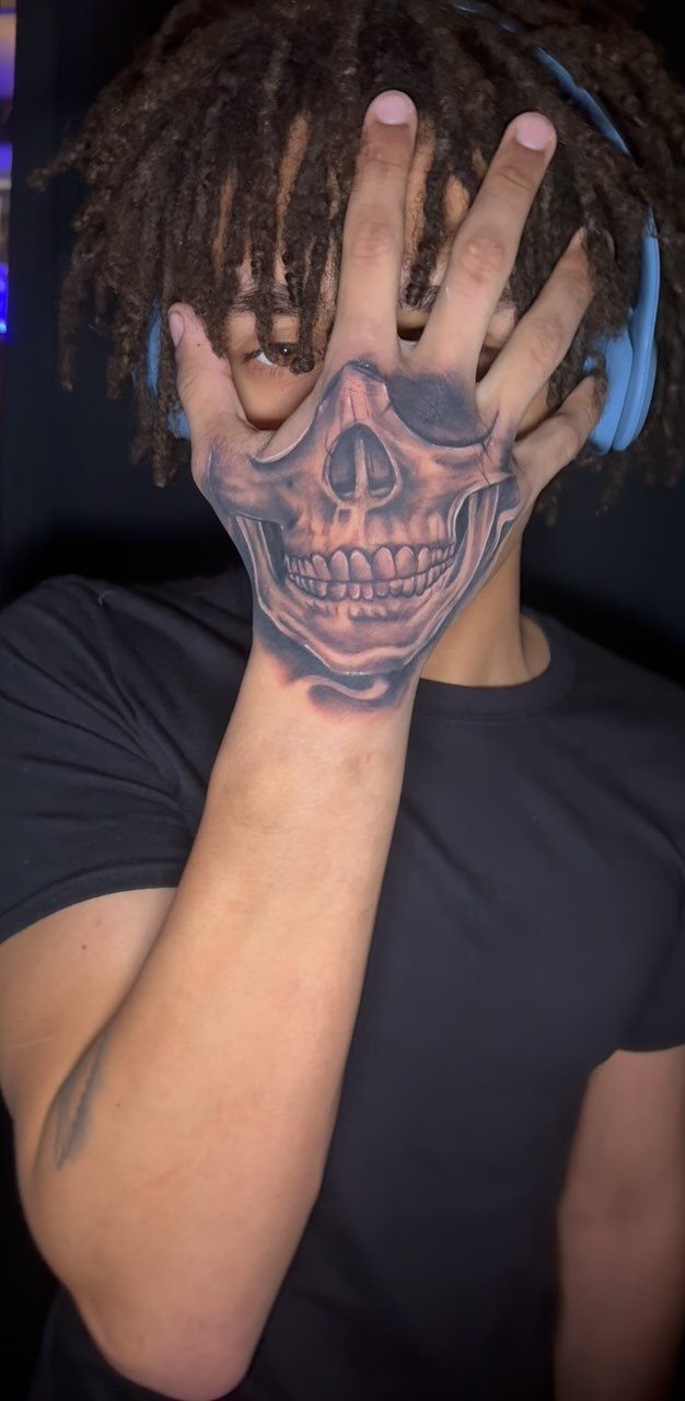 Detailed skull tattoo on the back of a man's hand covering his face.