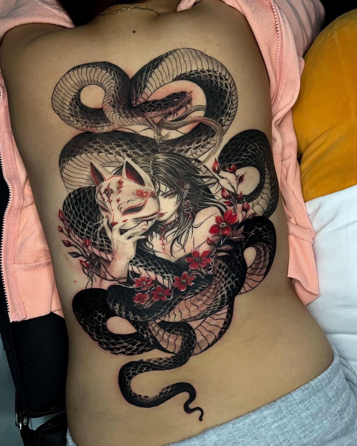 Large snake and kitsune mask tattoo on a woman's back with red flowers.