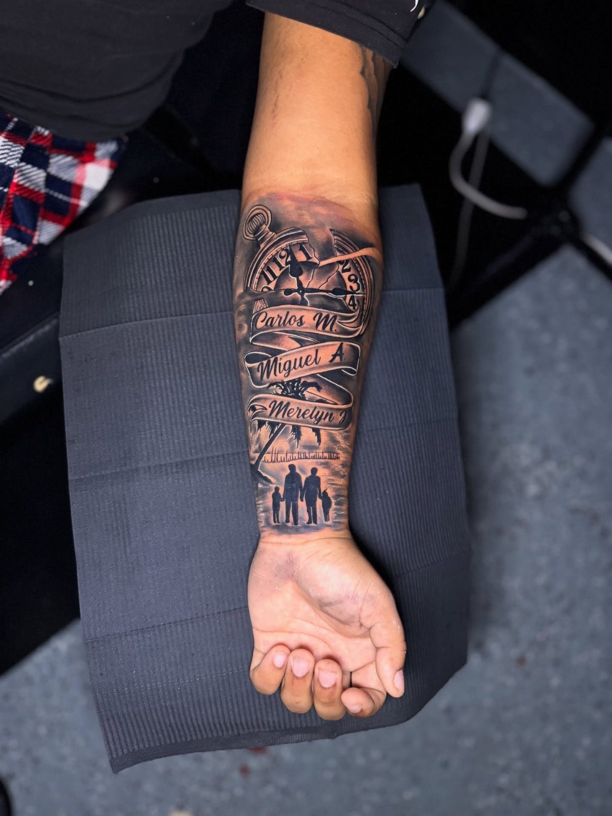 Lower arm tattoo featuring family silhouettes, names in script, and a cityscape.