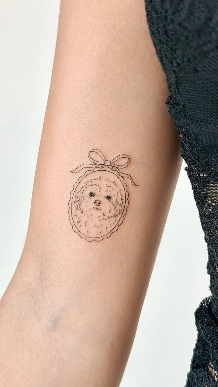 Small dog portrait tattoo inside an oval frame with a bow on a forearm.