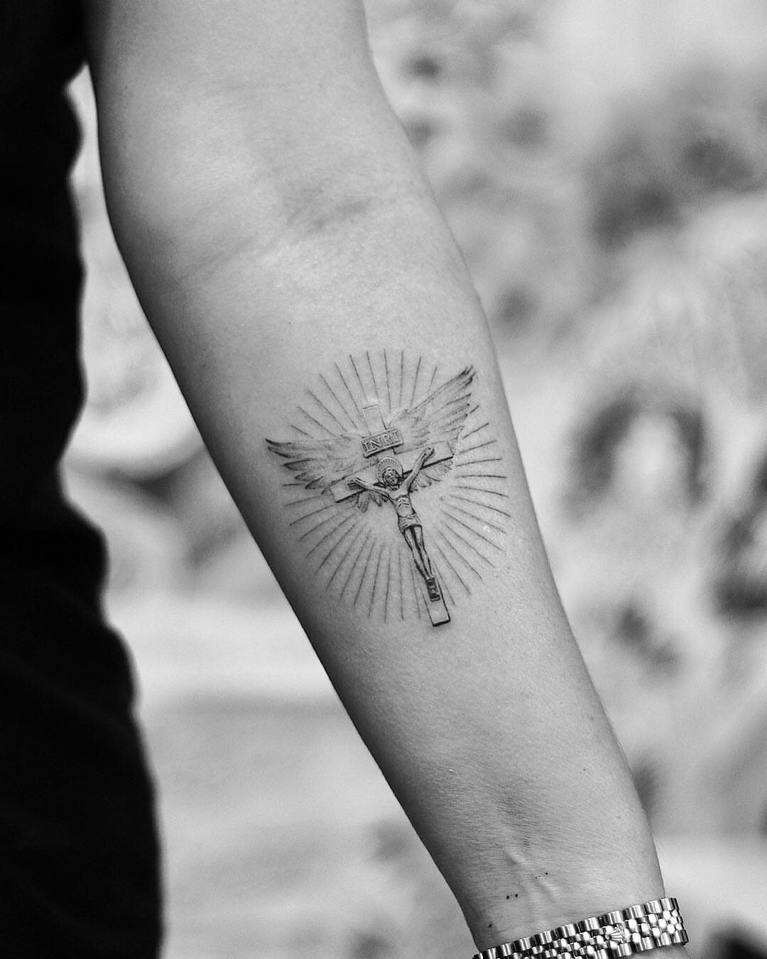 Crucifix tattoo with wings and radiating lines on a person's forearm.