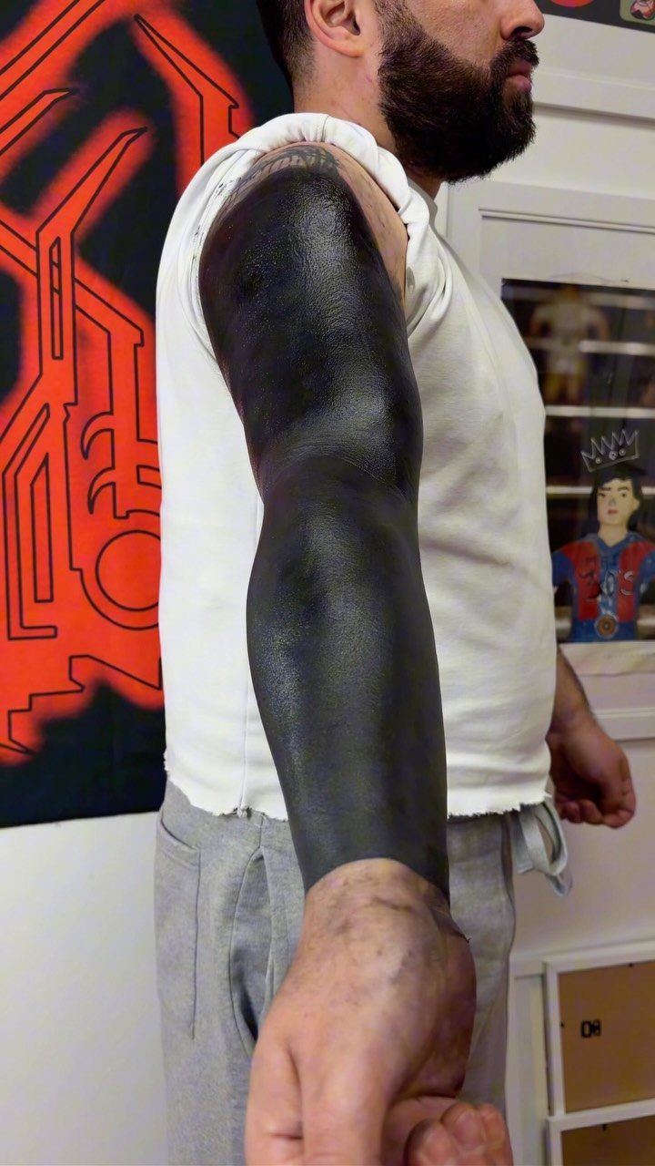 Man's arm covered in a solid black ink blackout sleeve tattoo.