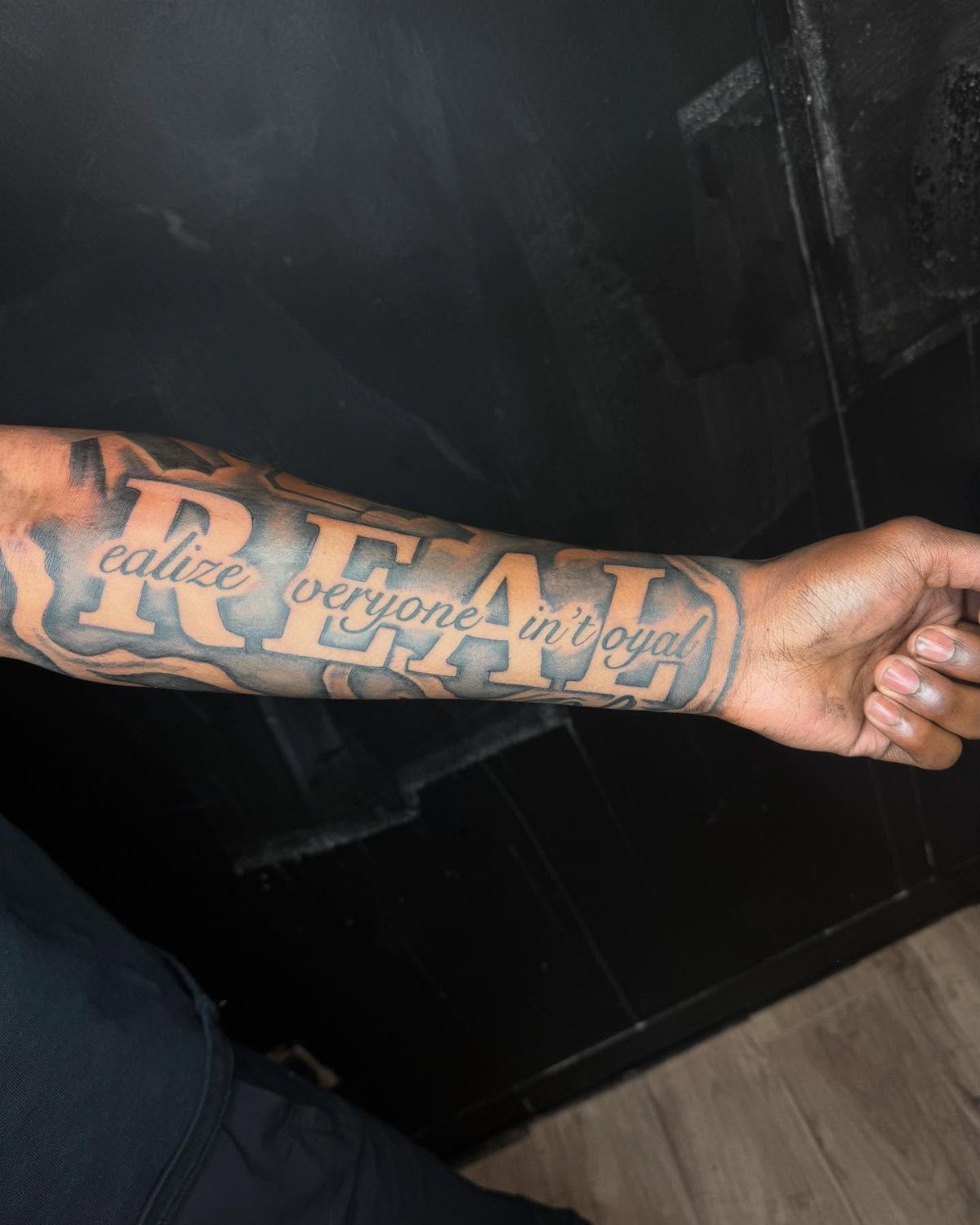 Forearm tattoo with the word REAL and a cursive quote in black ink