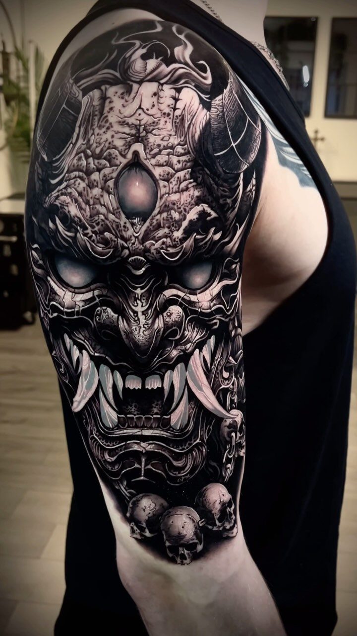 Demonic mask tattoo with horns and skulls on a man's upper arm sleeve.