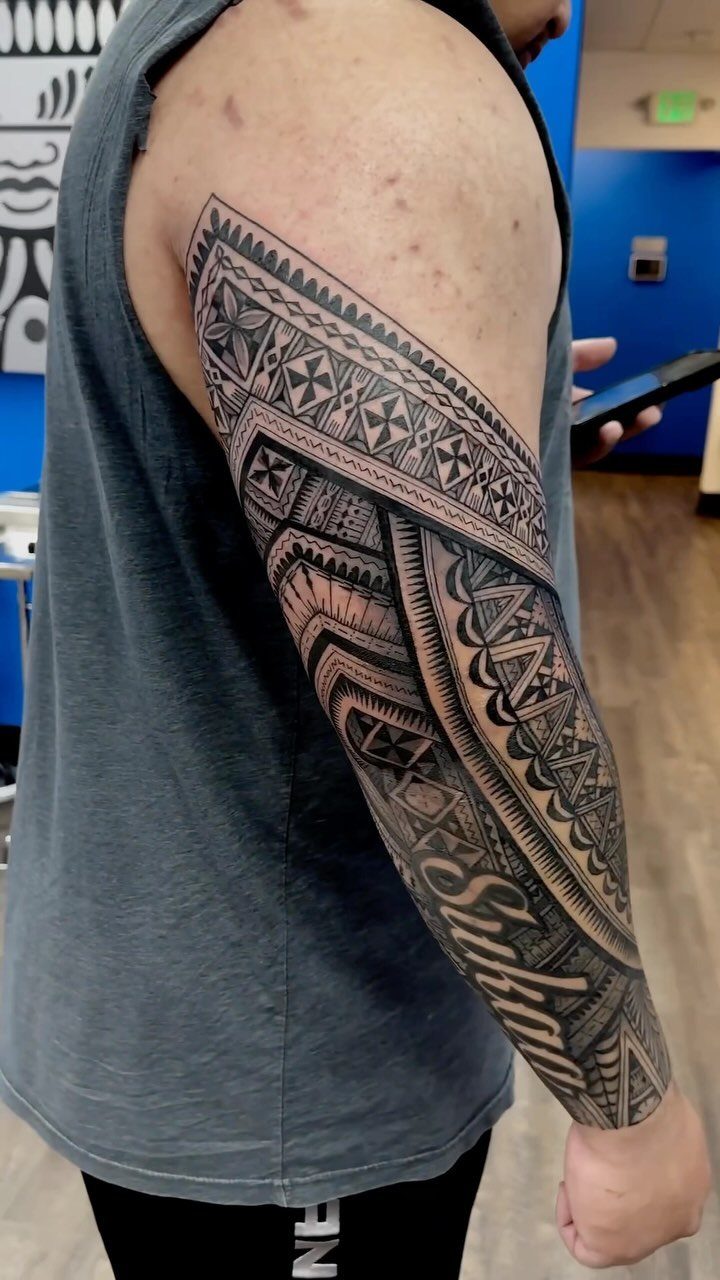Man's arm with a full sleeve tribal tattoo featuring black geometric patterns and lettering.