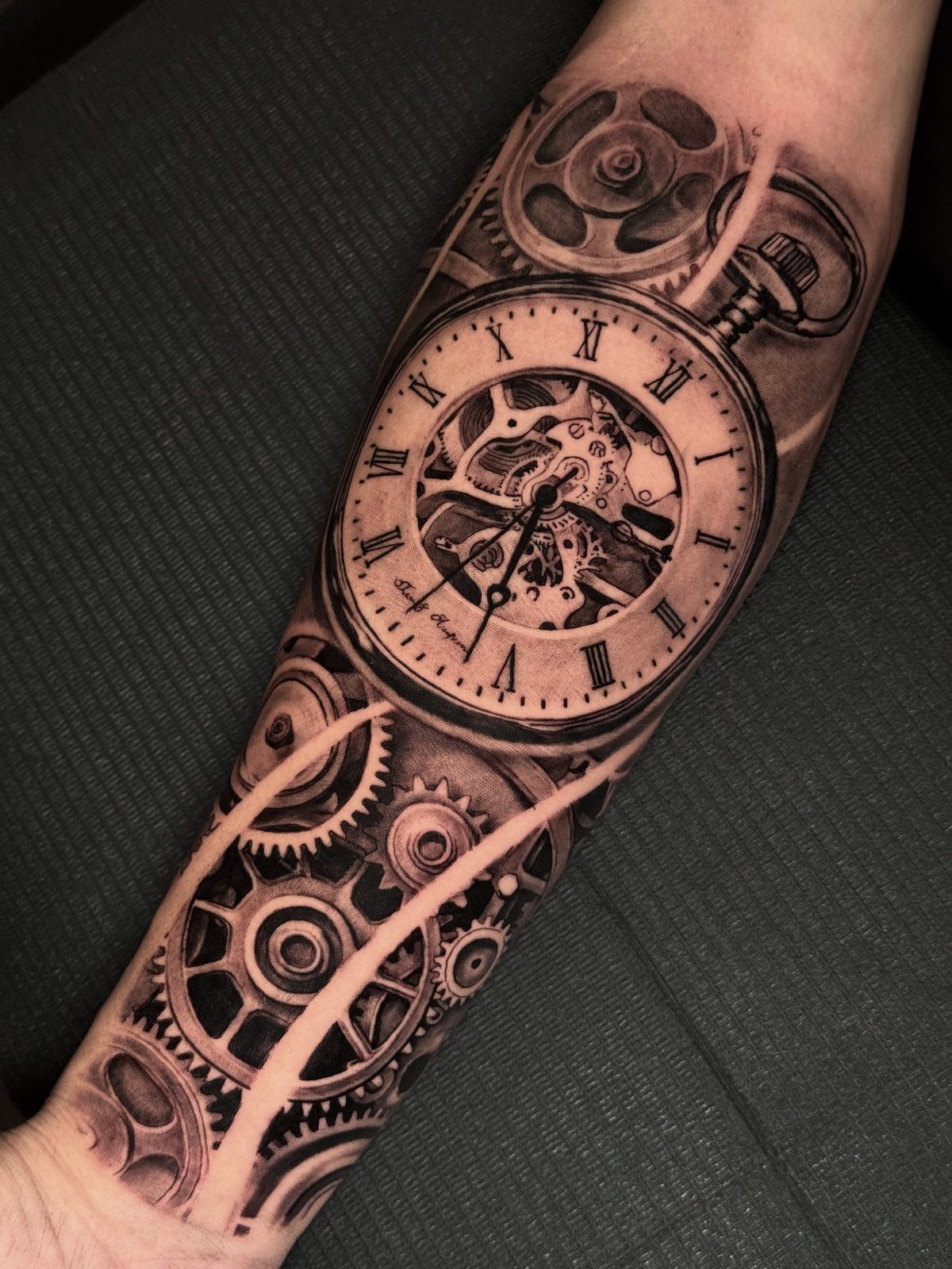 Detailed black and grey tattoo of a pocket watch and gears on a forearm.