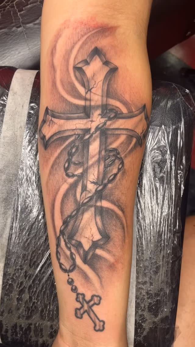 Large cracked stone cross tattoo with a rosary chain on a person's forearm.