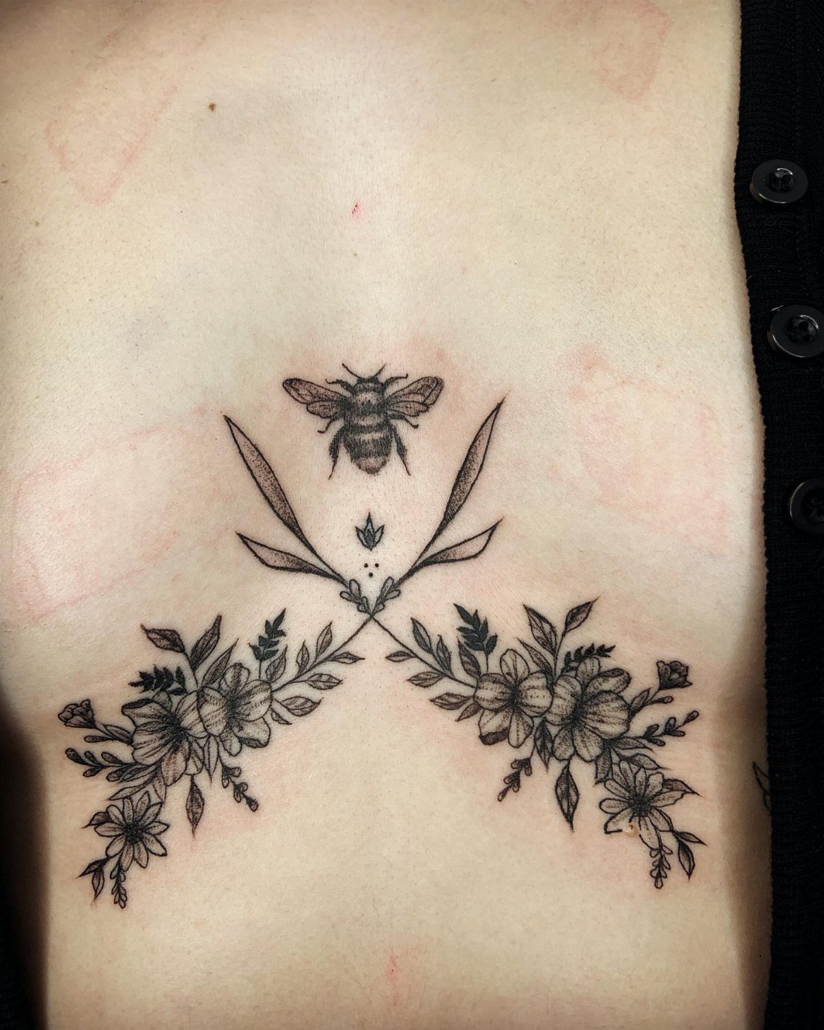 Bee and floral tattoo centered on a person's chest between the breasts.