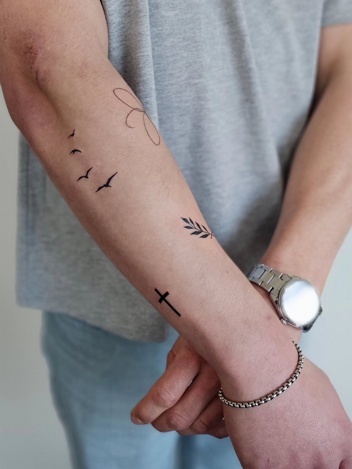 Forearm with minimalist black ink tattoos of birds, a leaf, and a cross.