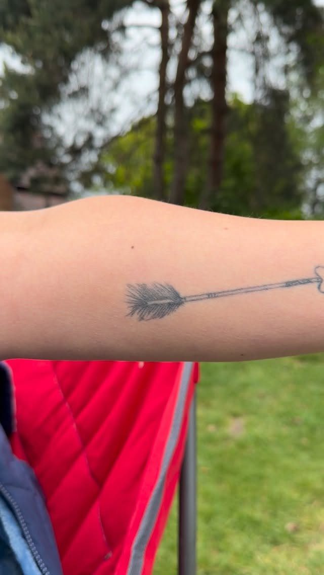 Small black arrow tattoo on a person's forearm in an outdoor setting.