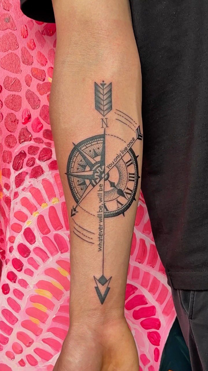 Compass tattoo with a vertical arrow and script text on a man's forearm.