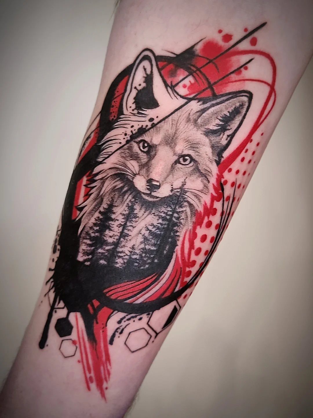 Fox head tattoo on a forearm with red abstract elements and forest silhouette.