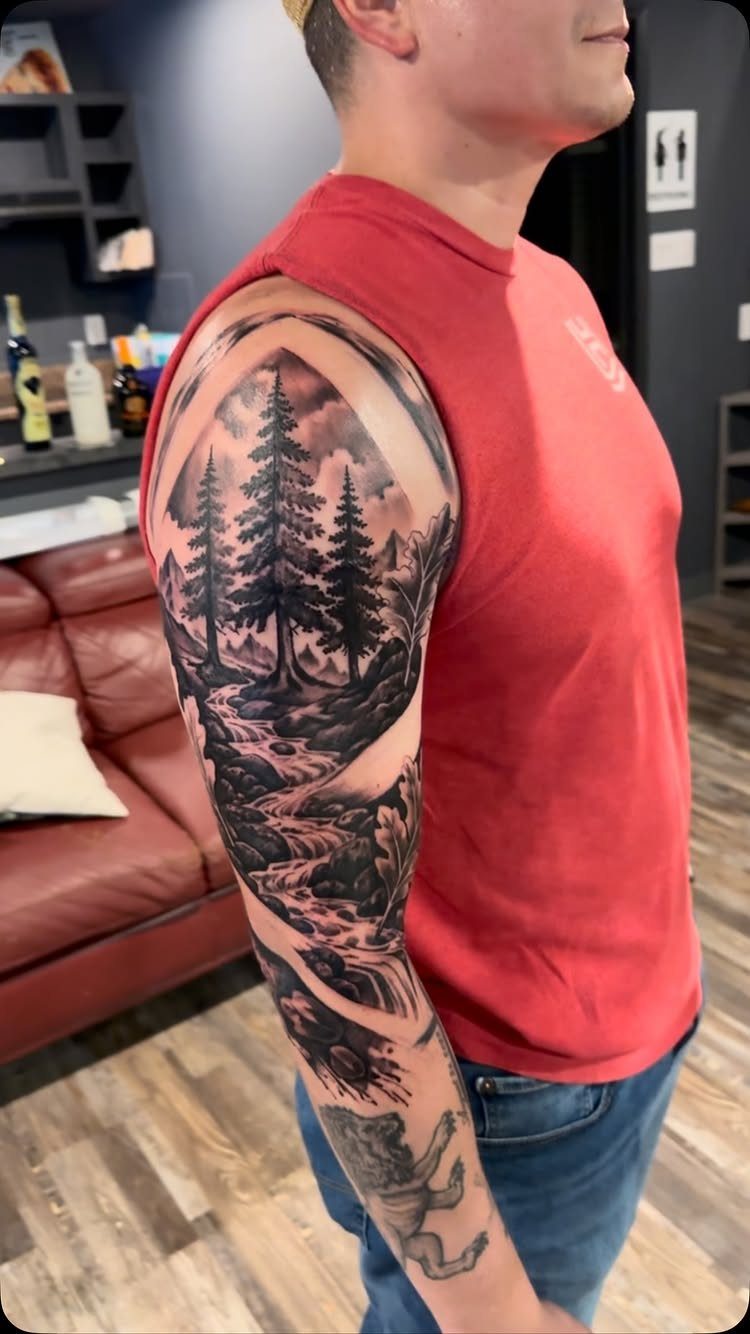 Man's arm with a detailed black and grey mountain and pine tree landscape tattoo.